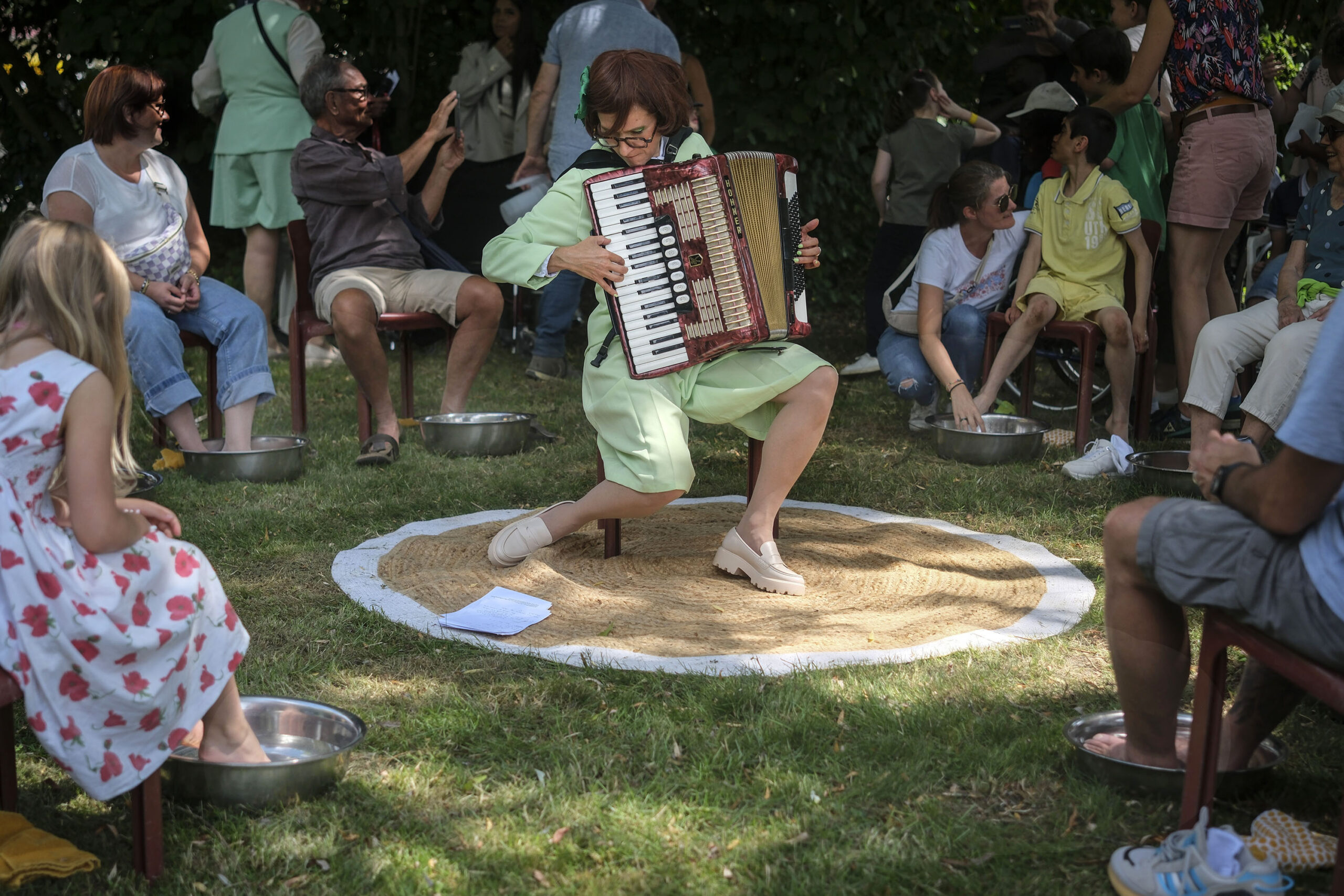 Cie ON-OFF-Plaines Sante-5 Cécile Thircuir, artiste de la compagnie On-Off, joue de l’accordéon à l’ombre dans un espace vert, entourée de participants installés les pieds dans des bassines métalliques remplies d’eau.