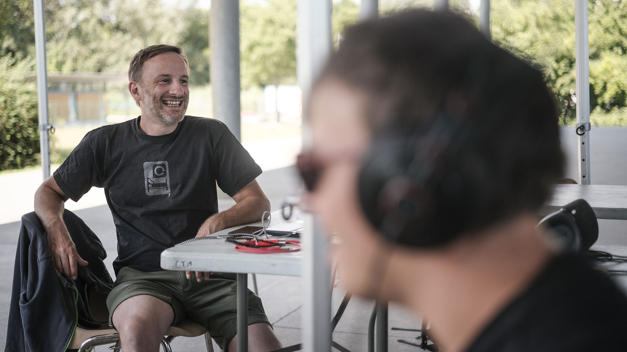 Otium_Plaines sante 47 L’artiste Matthieu Debliqui est assis, souriant. À droite de l’image, on distingue la silhouette floue d’une personne portant un casque audio